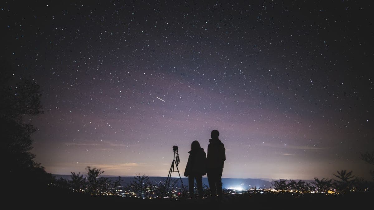 Two people watching a starry sky above distant town lights, a tripod beside them