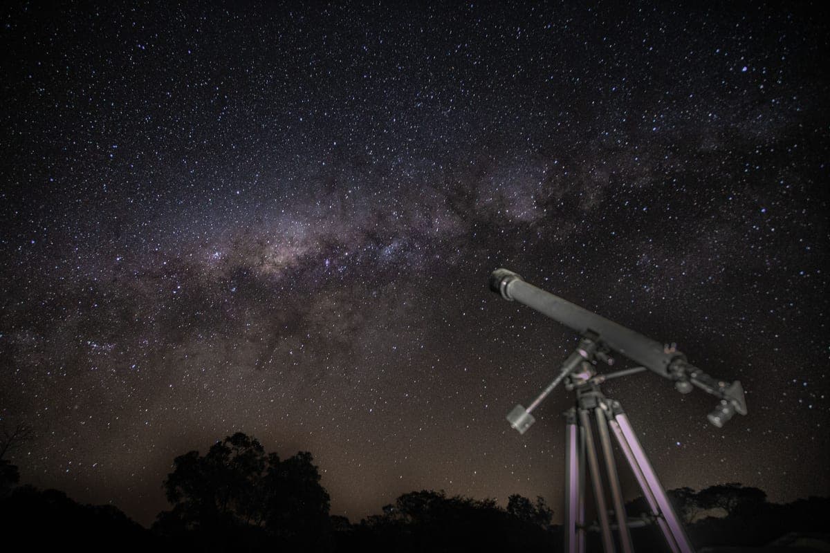 A telescope on a tripod pointed at the Milky Way over a dark treeline