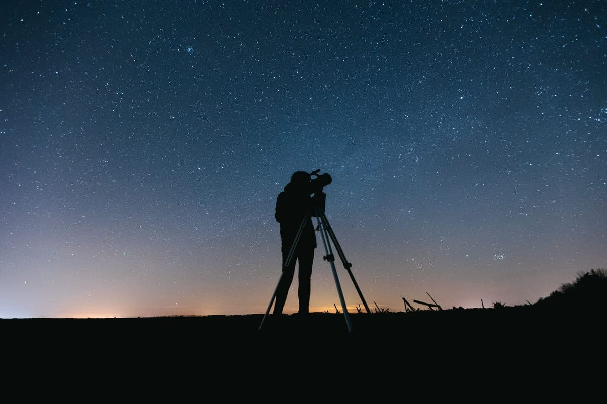 A person silhouetted beside a telescope under a starry sky